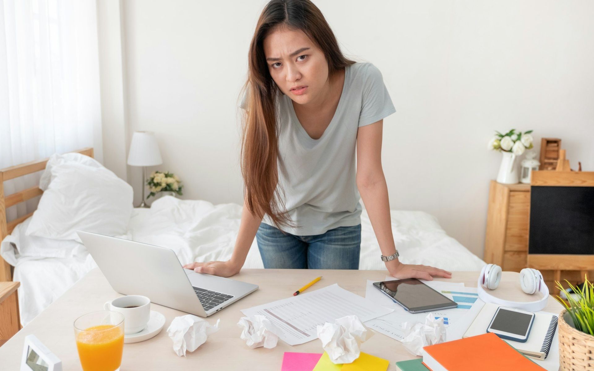 Jeune femme stressée dans une chambre en désordre, entourée de papiers froissés, d’un ordinateur et de tasses, illustrant l’impact du désordre sur le mental.